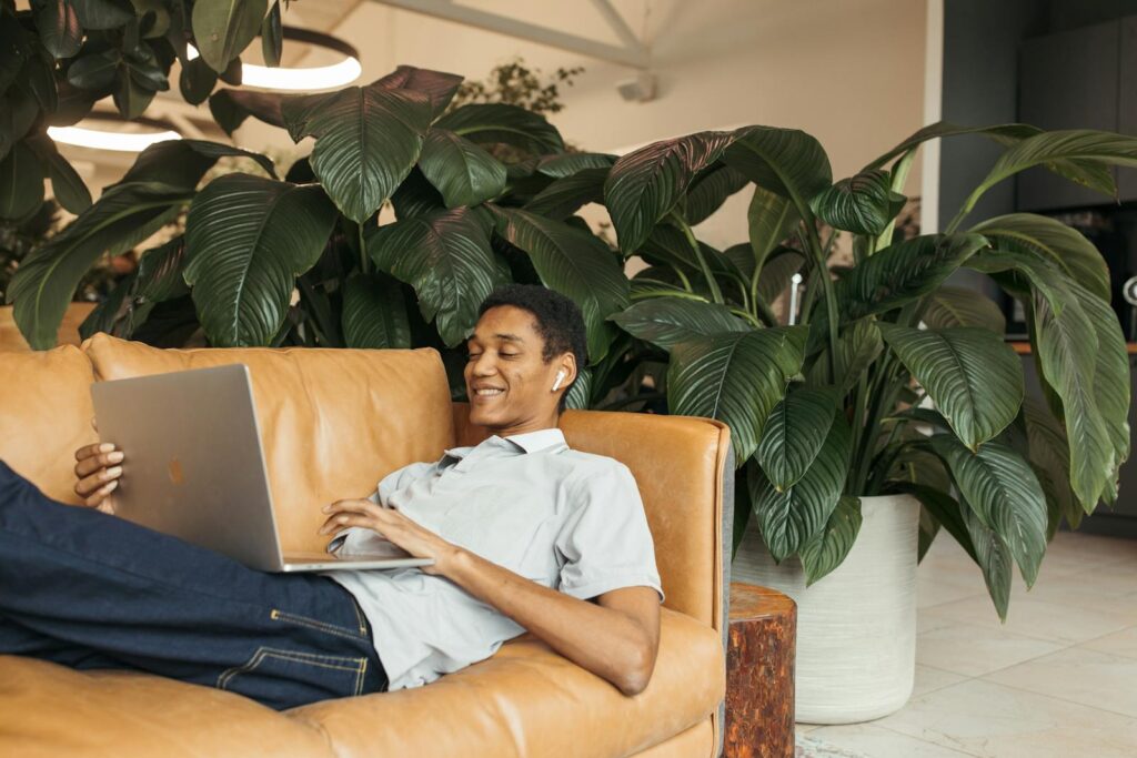 A man relaxing on a sofa using a laptop, surrounded by indoor plants.