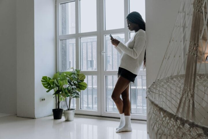 A young woman with braids uses her mobile phone in a minimalist room with plants and a hammock chair.